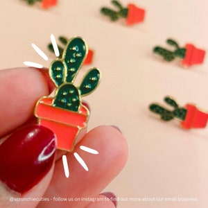 May include: A close-up of a green cactus enamel pin with a red pot. The pin is held in a hand with red nail polish. The background is a light pink.