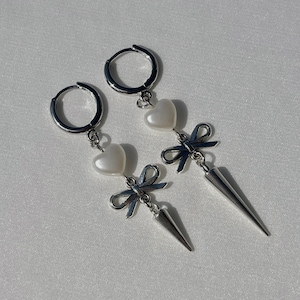 May include: Silver hoop earrings with dangling charms. Each earring has a white heart-shaped bead, a bow charm, and a silver spike pendant. The earrings are presented on a white surface.