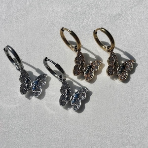 May include: Three pairs of hoop earrings with butterfly charms. The earrings are silver, gold, and rose gold. The butterflies are adorned with sparkling rhinestones.