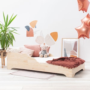 Könnte beinhalten: Ein helles Holz-Kleinkindbett mit einer braunen Decke und einem Teddybären auf der Matratze. Das Bett steht in einem Zimmer mit einer weißen Wand und einer Topfpflanze.