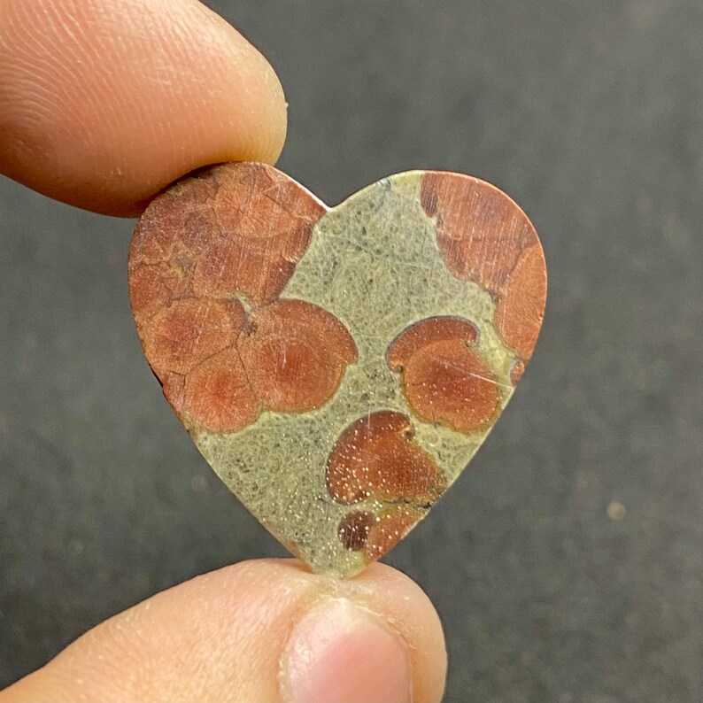 May include: Heart-shaped stone with a unique pattern. The stone features a mix of reddish-brown and light green colors. The reddish-brown areas have a textured, cracked appearance, while the green areas are smoother. The stone is held by a finger.