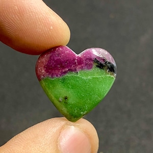 Op de afbeelding: Een hartvormige edelsteen met een levendige kleurencombinatie. De bovenste helft is diep magenta, overgaand in heldergroen aan de onderkant. De steen heeft een gepolijste, glanzende afwerking en wordt tussen twee vingers gehouden.