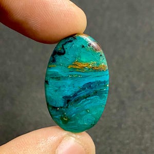 Op de afbeelding: Een ovaalvormige gepolijste edelsteen met een levendige mix van turquoise, blauwgroene en diepblauwe kleuren met opvallende zwarte aders en vlekken van aardebruine insluitingen over het gladde, glanzende oppervlak van de steen.