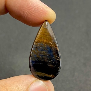 May include: A teardrop-shaped gemstone with a polished surface. The stone features a blend of colors, including gold, deep blue, and black, with a shimmering, reflective quality. The gemstone is held between two fingers against a dark background.