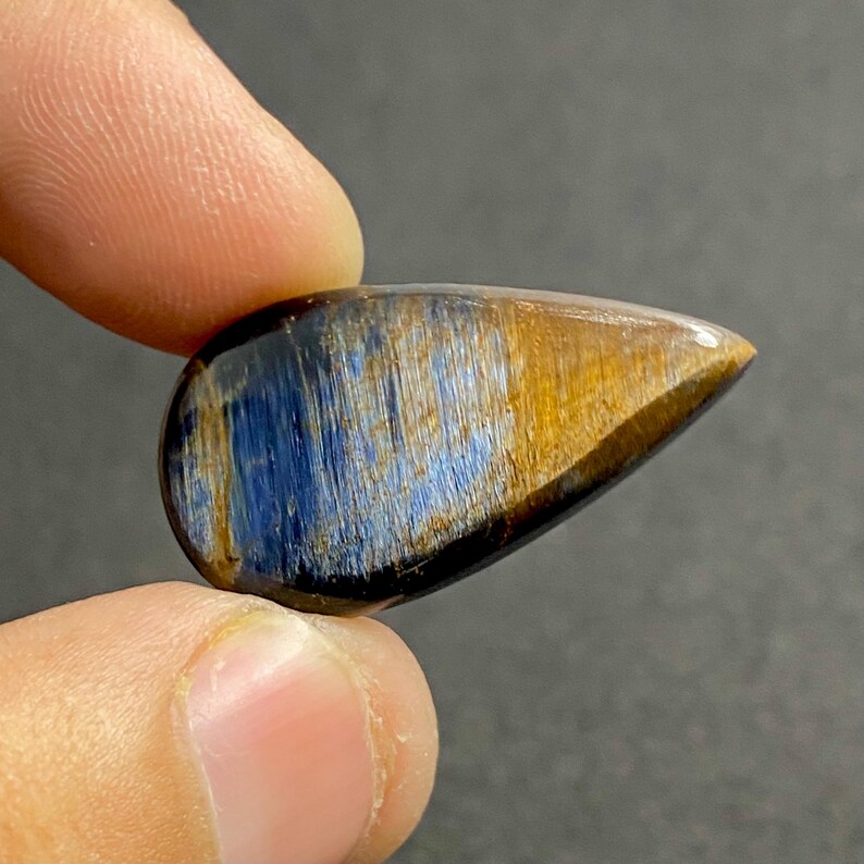 May include: A teardrop-shaped gemstone with a polished surface. The stone features a blend of blue, gold, and brown hues, with a chatoyant effect. The gemstone is held between two fingers, showcasing its natural beauty and unique color patterns.