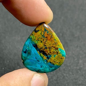 Könnte beinhalten: Ein tropfenförmiger Cabochon-Edelstein mit einer deutlichen Farbtrennung. Der obere Teil zeigt eine texturierte, gesprenkelte goldbraune und ockerfarbene Oberfläche, während der untere Bereich in ein leuchtendes, poliertes Türkisblau übergeht.