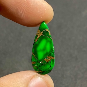 May include: A pear-shaped gemstone, predominantly green with gold-coloured veins. The polished surface catches the light, revealing natural patterns. The gemstone is held between two fingers, emphasising its size and shape.