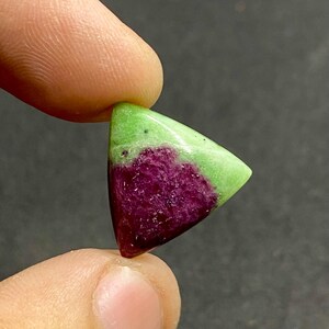 May include: A triangular gemstone with a vibrant color combination. The top portion is a bright green, transitioning to a deep magenta hue at the base. The stone is held between two fingers, showcasing its polished surface.
