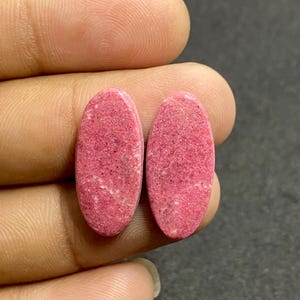 May include: Two oval-shaped, cabochon-cut gemstones with a vibrant, saturated pink color. The stones feature a textured, matte surface with subtle, lighter pink and white mineral inclusions distributed across the opaque, rounded faces.
