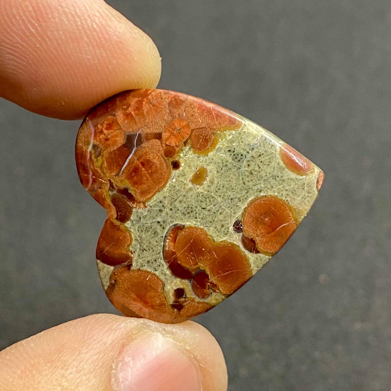 May include: Heart-shaped gemstone with a unique pattern. The stone features a mix of colors, including orange, red, and beige. The surface has a textured appearance with circular and irregular patterns. The gemstone is held between two fingers.
