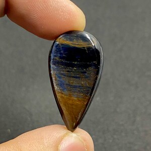 May include: A polished, teardrop-shaped gemstone with a striking pattern of blue and gold hues. The stone is held between two fingers, showcasing its glossy surface and intricate internal structure. The gem's colors shift and shimmer, creating a captivating visual effect.