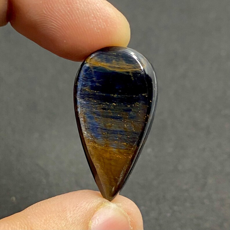 May include: A polished, teardrop-shaped gemstone with a striking pattern of blue and gold hues. The stone is held between two fingers, showcasing its glossy surface and intricate internal structure. The gem's colors shift and shimmer, creating a captivating visual effect.