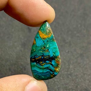 Op de afbeelding: Een traanvormige gepolijste edelsteen met een levendige mix van turquoise, blauwgroen en diepblauwe kleuren met duidelijke zwarte aders en vlekken van goudbruine matrix over het gehele oppervlak, met een gladde en glanzende afwerking.