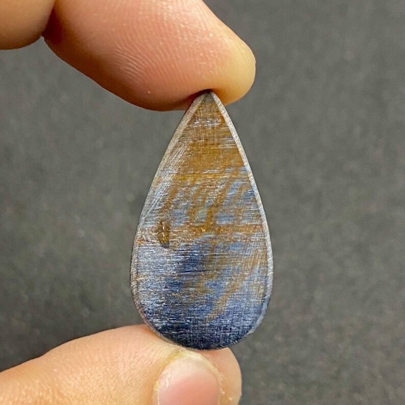 May include: A teardrop-shaped gemstone with a polished surface. The stone features a blend of blue and gold hues, with visible linear patterns. Held between fingers, the gem's shape is elongated and symmetrical, suitable for jewelry.