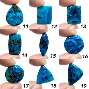 Op de afbeelding: Een verzameling van negen gepolijste edelstenen in verschillende vormen, waaronder druppel, rechthoekig en driehoekig. De stenen vertonen een levendige blauwe kleur met donkerdere blauwe en bruine adering. Elke steen wordt tussen vingers gehouden.