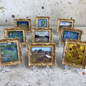 May include: A collection of nine miniature paintings in ornate gold-colored frames, each displayed on a small clear plastic stand. The artworks feature various landscape and still life scenes, including sunflowers, portraits, and fields in vibrant colors.