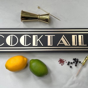 以下が含まれることがあります： 「COCKTAILS」の文字が金色で書かれた黒と金の看板、金色のジガー、レモン、ライム、ベリー、カクテルピック。看板は長方形です。