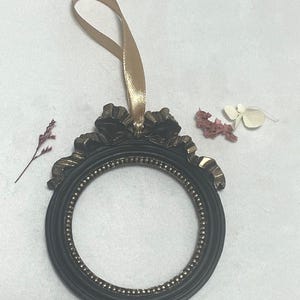 Può includere: Una cornice ovale nera e dorata con un nastro dorato per appenderla. La cornice ha una parte superiore decorativa e un bordo interno perlato. Fiori secchi sono accanto alla cornice.