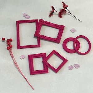 May include: A collection of small, bright pink picture frames in various shapes, including rectangular and square, are arranged on a white surface. Decorative dried flowers and petals are scattered around the frames, adding a touch of elegance.