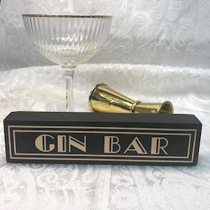 Op de afbeelding: Een zwart houten bord met de woorden "GIN BAR" in gouden letters, een coupeglas met gouden rand en een gouden jigger. Het bord heeft een gouden rand en een lettertype in art deco-stijl.