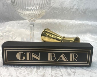 May include: A black wooden sign with the words "GIN BAR" in gold lettering, a gold-rimmed coupe glass, and a gold jigger. The sign has a gold border and art deco style font.