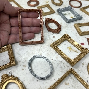 May include: A collection of miniature picture frames in various shapes, sizes, and metallic finishes including gold, copper, and silver. The frames feature intricate embossed detailing along the borders, with some rectangular and some oval in design.