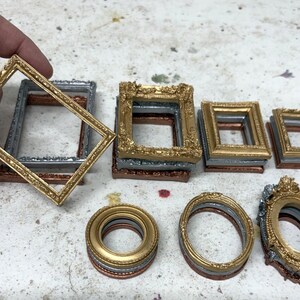 May include: A collection of miniature picture frames in various shapes and sizes, including rectangular, oval, and ornate styles. The frames are finished in metallic gold, silver, and copper tones, stacked in small groups on a speckled surface.