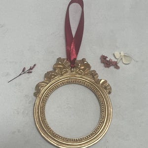 May include: A gold-coloured, ornate picture frame with a burgundy ribbon for hanging. The frame is round and has a decorative, scalloped edge.