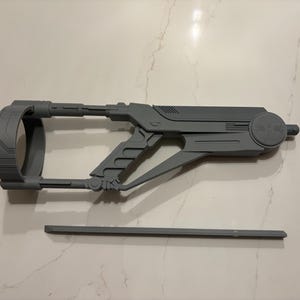 Könnte beinhalten: Eine graue Requisitenpistole im futuristischen Stil mit einem kreisförmigen Element und einem Griff mit triggerartiger Form. Ein separates, dünnes, rechteckiges Teil befindet sich darunter. Das Objekt besteht aus einem festen Material.