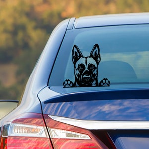 May include: Black vinyl car decal featuring a German Shepherd dog design. The dog's head and paws are visible, giving the impression it's peeking over the edge. The decal is applied to the rear window of a blue car.