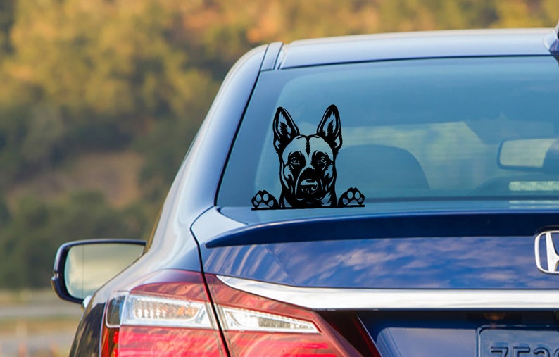 May include: Black vinyl car decal featuring a German Shepherd dog design. The dog's head and paws are visible, giving the impression it's peeking over the edge. The decal is applied to the rear window of a blue car.