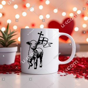 May include: White ceramic mug with a black and white illustration of a lamb carrying a cross. The mug is on a red heart-shaped confetti background.