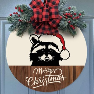 May include: A round wooden door hanger with a black and white raccoon wearing a Santa hat. The top half of the door hanger is cream colored and the bottom half is a dark wood. The text "Merry Christmas" is written in white on the wood. A red and black buffalo plaid bow with green pine sprigs and red berries is attached to the top of the door hanger.