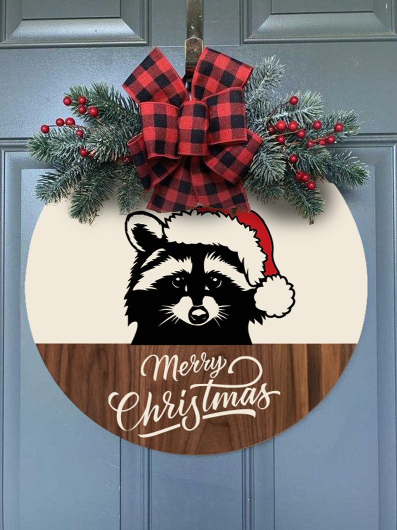 May include: A round wooden door hanger with a black and white raccoon wearing a Santa hat. The top half of the door hanger is cream colored and the bottom half is a dark wood. The text "Merry Christmas" is written in white on the wood. A red and black buffalo plaid bow with green pine sprigs and red berries is attached to the top of the door hanger.