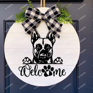May include: A round, white wooden welcome sign featuring a black and white dog graphic. The sign has a black and white buffalo check bow and greenery at the top. The word "Welcome" is written in script below the dog.
