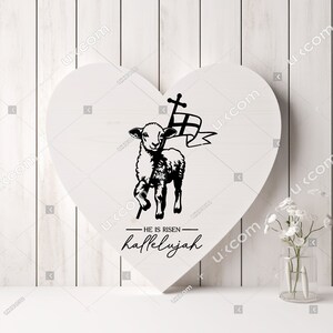 May include: A white heart-shaped wooden sign with a black silhouette of a lamb carrying a cross and a banner. The text "HE IS RISEN - hallelujah" is written below the lamb.