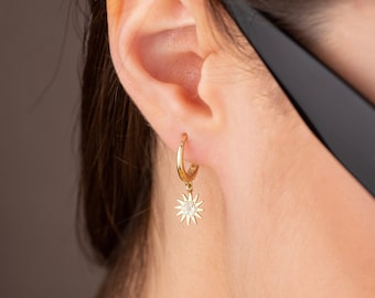 Peut inclure: Boucle d'oreille créole dorée avec un charm en forme d'éclat de soleil. Le charm a une pierre centrale et des pointes rayonnantes. Le mot "VOGUE" est imprimé sur les lunettes de soleil noires en arrière-plan.