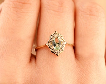 May include: A gold ring featuring a central oval-cut light green gemstone surrounded by a halo of small clear stones set in a scalloped, pointed frame. The thin gold band supports the decorative setting, which has a distinct pointed shape.