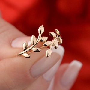 May include: A gold-coloured ring featuring a delicate leaf design. The ring has two branches of leaves, each with multiple leaves. The leaves have a smooth, polished finish. The ring is a piece of jewellery.