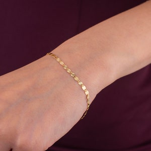 K&ouml;nnte beinhalten: Ein zartes goldenes Armband mit einer Reihe kleiner, flacher, kreisf&ouml;rmiger Anh&auml;nger, die miteinander verbunden sind. Das Armband wird am Handgelenk getragen und zeigt sein schlichtes, aber elegantes Design. Die Goldfarbe sorgt f&uuml;r eine warme, klassische &Auml;sthetik.