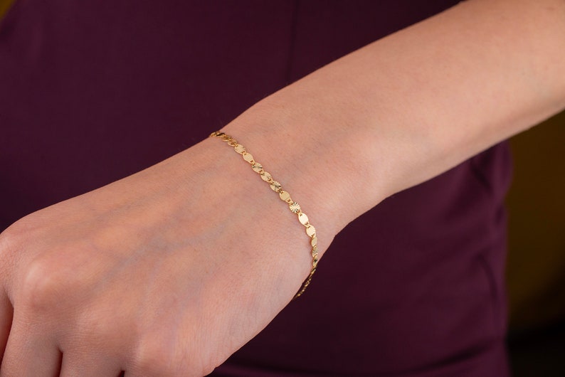 K&ouml;nnte beinhalten: Ein zartes goldenes Armband mit einer Reihe kleiner, flacher, kreisf&ouml;rmiger Anh&auml;nger, die miteinander verbunden sind. Das Armband wird am Handgelenk getragen und zeigt sein schlichtes, aber elegantes Design. Die Goldfarbe sorgt f&uuml;r eine warme, klassische &Auml;sthetik.