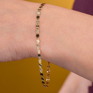 K&ouml;nnte beinhalten: Ein zartes goldenes Armband mit einem sich wiederholenden Muster aus kleinen, runden und sternf&ouml;rmigen Anh&auml;ngern. Das Armband wird am Handgelenk einer Person mit gelbem Hintergrund gezeigt.