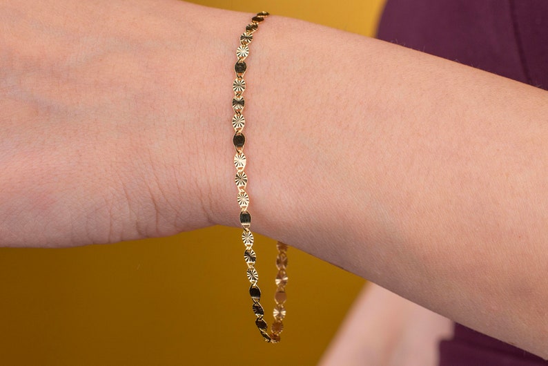 K&ouml;nnte beinhalten: Ein zartes goldenes Armband mit einem sich wiederholenden Muster aus kleinen, runden und sternf&ouml;rmigen Anh&auml;ngern. Das Armband wird am Handgelenk einer Person mit gelbem Hintergrund gezeigt.