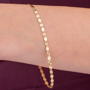 K&ouml;nnte beinhalten: Ein zartes goldenes Armband mit einem sich wiederholenden Muster aus ovalen und sternf&ouml;rmigen Gliedern. Das Armband wird an einem Arm gezeigt, wobei die warme Goldfarbe mit dem Hautton kontrastiert. Das Schmuckst&uuml;ck ist schlicht und elegant.