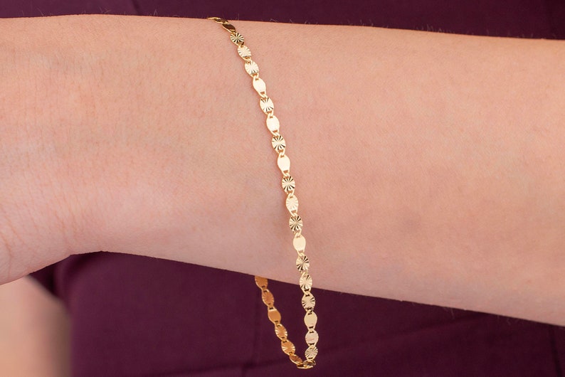 K&ouml;nnte beinhalten: Ein zartes goldenes Armband mit einem sich wiederholenden Muster aus ovalen und sternf&ouml;rmigen Gliedern. Das Armband wird an einem Arm gezeigt, wobei die warme Goldfarbe mit dem Hautton kontrastiert. Das Schmuckst&uuml;ck ist schlicht und elegant.