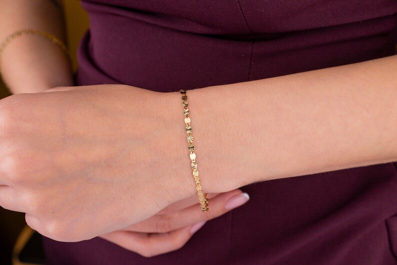 K&ouml;nnte beinhalten: Ein zartes goldenes Armband mit einem sich wiederholenden Muster aus kleinen, flachen, oval geformten Gliedern. Das Armband wird an einem Handgelenk vor einem dunkelburgunderfarbenen Hintergrund getragen. Das Armband scheint aus Gold zu sein.