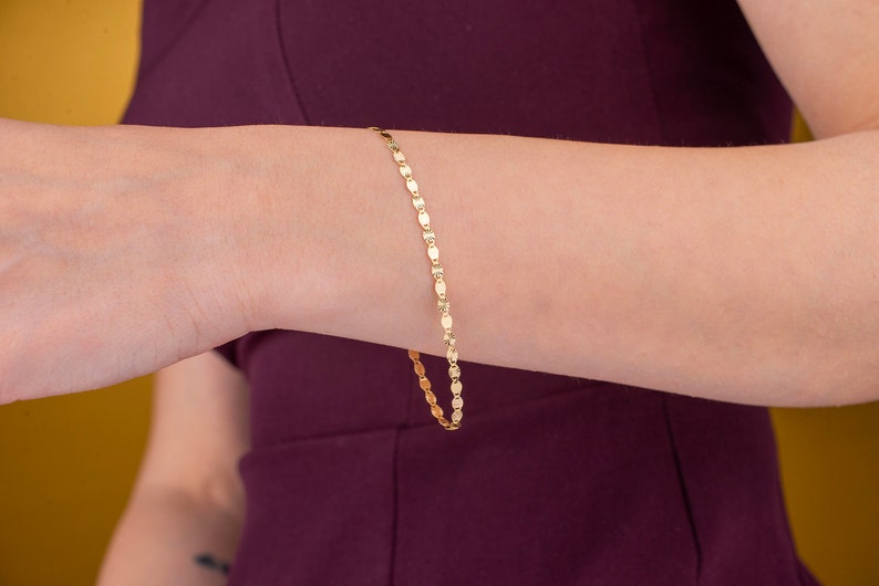 K&ouml;nnte beinhalten: Ein zartes goldenes Armband mit einem ovalen Kettendesign. Das Armband wird am Handgelenk getragen, und die Goldfarbe kontrastiert mit dem dunklen Lila des &Auml;rmels. Der Hintergrund ist ein warmes Gelb.