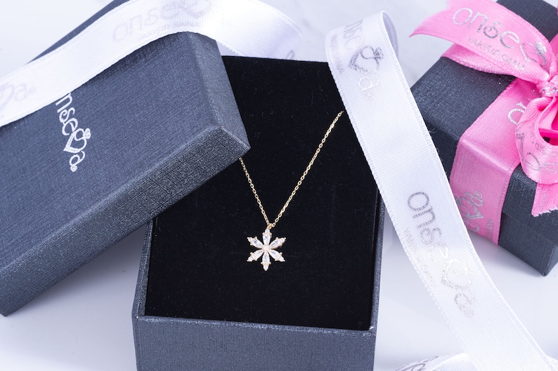 Puede incluir: Un collar dorado con un colgante de copo de nieve, presentado en una caja de regalo negra. El colgante est&aacute; adornado con cristales transparentes. Cinta blanca con texto plateado y una caja de regalo rosa tambi&eacute;n visibles.