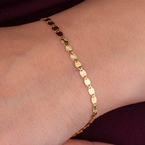 K&ouml;nnte beinhalten: Ein zartes goldenes Armband mit einer Reihe kleiner, flacher, kreisf&ouml;rmiger Anh&auml;nger. Einige Anh&auml;nger haben ein Sternmuster. Das Armband wird an einem Handgelenk vor burgunderrotem Hintergrund getragen.