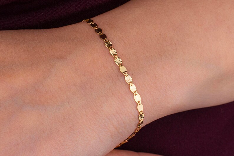 K&ouml;nnte beinhalten: Ein zartes goldenes Armband mit einer Reihe kleiner, flacher, kreisf&ouml;rmiger Anh&auml;nger. Einige Anh&auml;nger haben ein Sternmuster. Das Armband wird an einem Handgelenk vor burgunderrotem Hintergrund getragen.